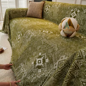 May include: An olive green sofa cover featuring a white geometric pattern. The cover is draped over a sofa, with a matching cushion and a decorative plush toy. The design includes a fringed edge for added texture.