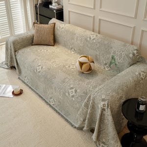 May include: A light grey sofa cover with a geometric pattern, draped over a sofa. A brown cushion and a decorative ball are on the sofa. A small side table and a cup of coffee are also visible.