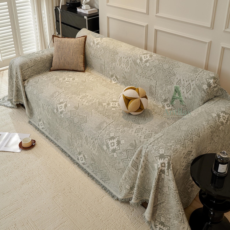 May include: A light grey sofa cover with a geometric pattern, draped over a sofa. A brown cushion and a decorative ball are on the sofa. A small side table and a cup of coffee are also visible.