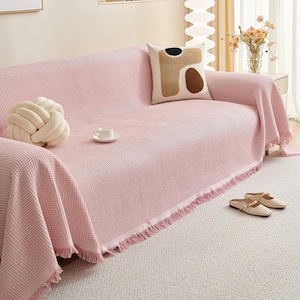 May include: A pink waffle-weave sofa cover with fringe trim, draped over a sofa. A decorative knot pillow and a patterned pillow rest on the sofa. A white teacup and saucer sit on the cover. Beige slippers are on the floor.