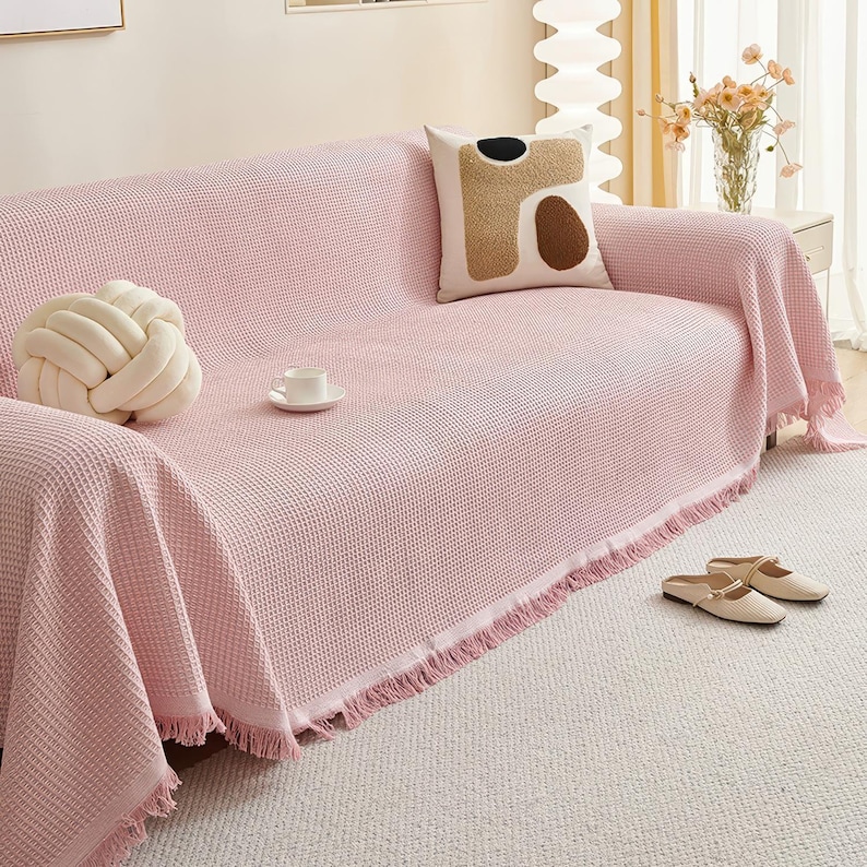 May include: A pink waffle-weave sofa cover with fringe trim, draped over a sofa. A decorative knot pillow and a patterned pillow rest on the sofa. A white teacup and saucer sit on the cover. Beige slippers are on the floor.