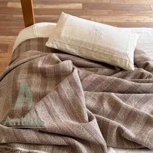 Pu&ograve; includere: Una coperta a righe marrone e beige con dettagli a frange &egrave; drappeggiata su un letto. Un cuscino bianco con finiture in pizzo e dettagli ricamati poggia sopra. Il letto ha un lenzuolo bianco.