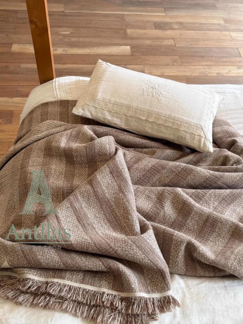 Pu&ograve; includere: Una coperta a righe marrone e beige con dettagli a frange &egrave; drappeggiata su un letto. Un cuscino bianco con finiture in pizzo e dettagli ricamati poggia sopra. Il letto ha un lenzuolo bianco.