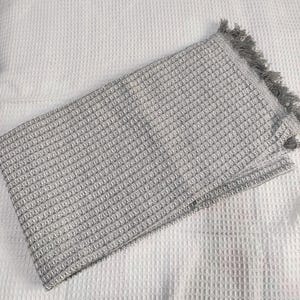May include: A folded gray waffle-weave blanket with a fringed edge. The blanket has a textured pattern and is made of a soft material. The blanket is folded on a white textured surface.