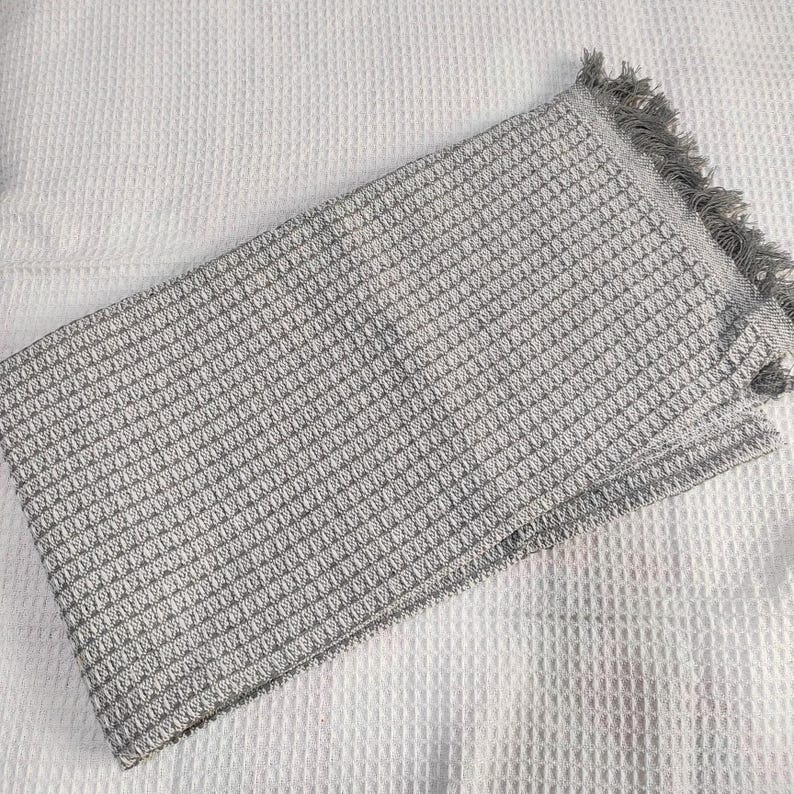May include: A folded gray waffle-weave blanket with a fringed edge. The blanket has a textured pattern and is made of a soft material. The blanket is folded on a white textured surface.