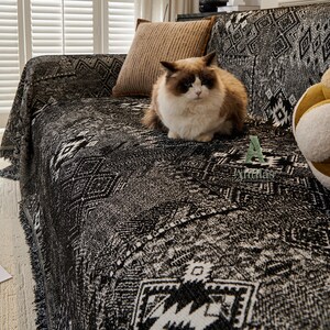 May include: A black and white patterned sofa throw blanket with fringe, draped over a sofa. A brown and white cat sits on the blanket. A brown cushion rests on the sofa. The blanket has a geometric design.