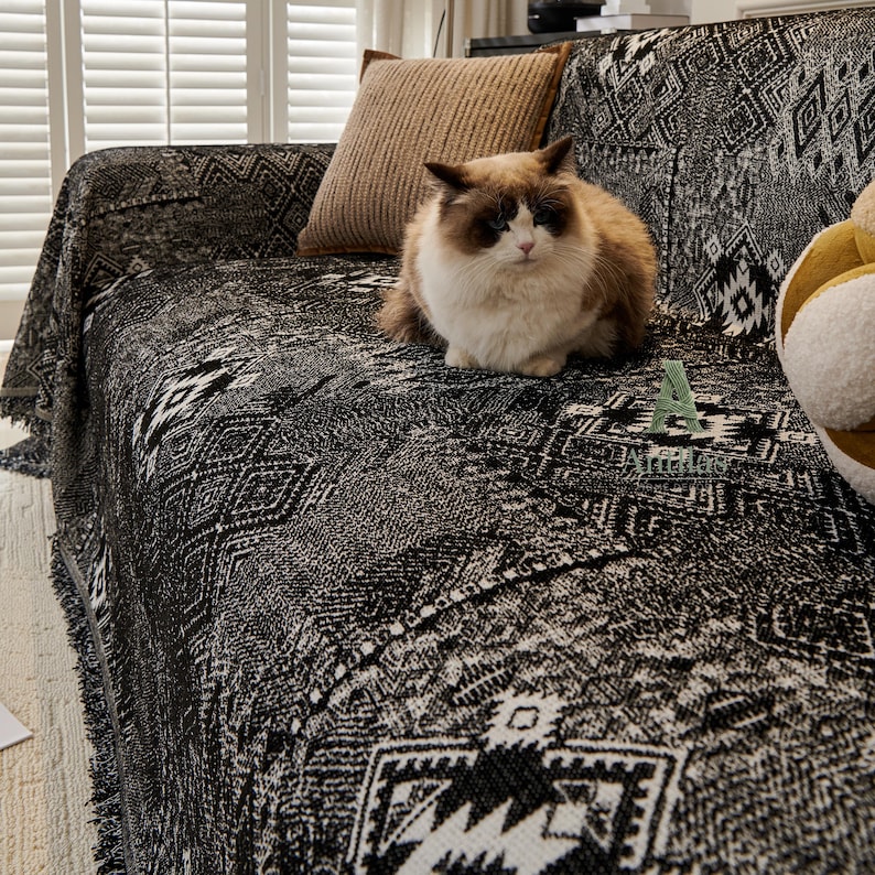 May include: A black and white patterned sofa throw blanket with fringe, draped over a sofa. A brown and white cat sits on the blanket. A brown cushion rests on the sofa. The blanket has a geometric design.