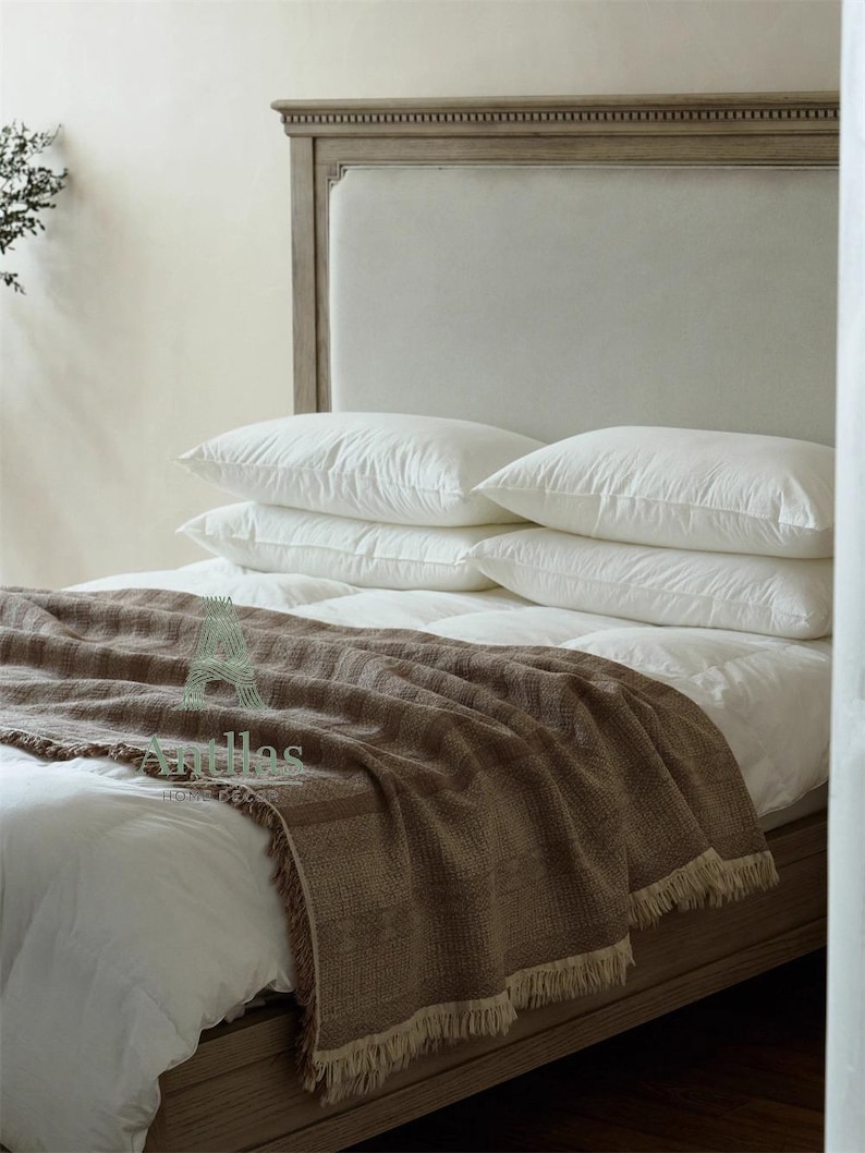 Pu&ograve; includere: Un letto con struttura e testiera in legno chiaro. Il letto &egrave; fatto con biancheria da letto bianca, compresi cuscini e un piumino. Una coperta marrone intrecciata con frange &egrave; drappeggiata sui piedi del letto. L'estetica generale &egrave; neutra e accogliente.