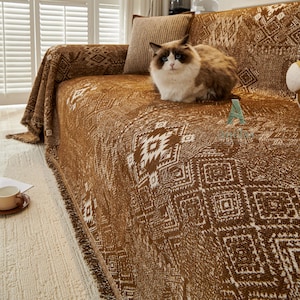 May include: A brown sofa throw with a white geometric design. The throw is draped over a sofa, with a fluffy cat on top. The throw has a fringed edge. A brown cushion is behind the cat.