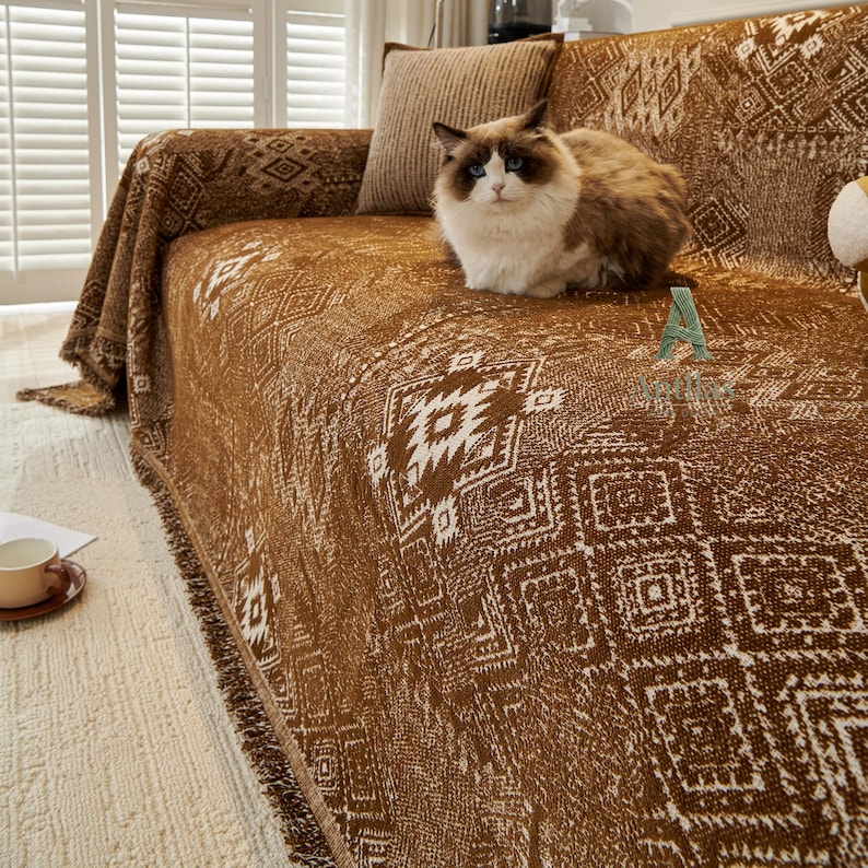 May include: A brown sofa throw with a white geometric design. The throw is draped over a sofa, with a fluffy cat on top. The throw has a fringed edge. A brown cushion is behind the cat.