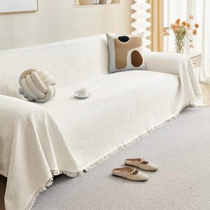 May include: A white waffle-weave sofa cover with fringe detail, draped over a sofa. A decorative knot pillow, a patterned pillow, and a teacup are on the sofa. Beige slip-on shoes rest on the gray carpet. A vase of flowers is in the background.