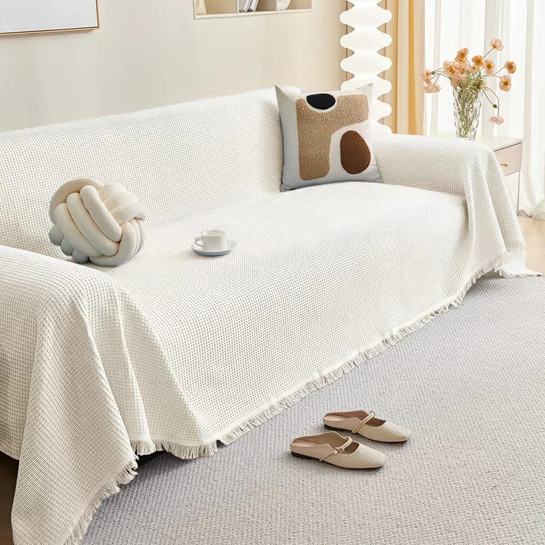 May include: A white waffle-weave sofa cover with fringe detail, draped over a sofa. A decorative knot pillow, a patterned pillow, and a teacup are on the sofa. Beige slip-on shoes rest on the gray carpet. A vase of flowers is in the background.