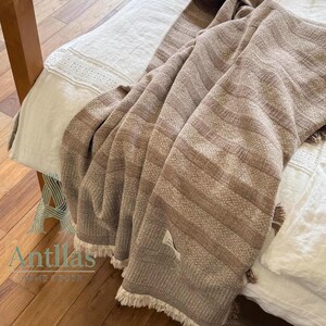 Pu&ograve; includere: Una coperta tessuta marrone e beige con un motivo a righe e frange. La coperta &egrave; drappeggiata su un letto con lenzuola bianche e un telaio in legno. L'etichetta della coperta recita "Antillas Home Decor".
