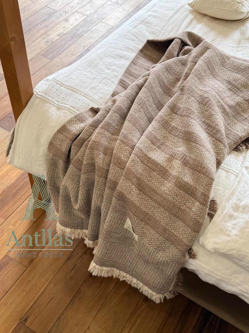 Pu&ograve; includere: Una coperta tessuta marrone e beige con un motivo a righe e frange. La coperta &egrave; drappeggiata su un letto con lenzuola bianche e un telaio in legno. L'etichetta della coperta recita "Antillas Home Decor".