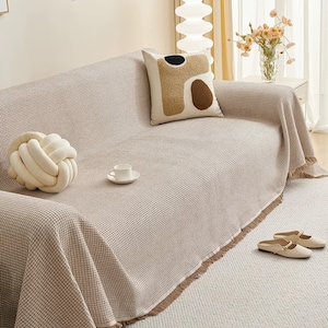 May include: A beige waffle-weave sofa cover with a fringed edge, draped over a sofa. Decorative pillows and a knotted cushion are on the sofa. A small teacup and saucer sit on the cover, and a pair of beige shoes are on the floor.