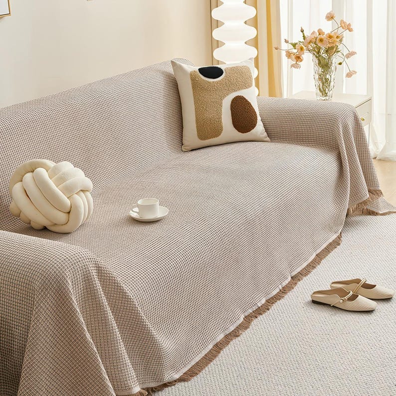 May include: A beige waffle-weave sofa cover with a fringed edge, draped over a sofa. Decorative pillows and a knotted cushion are on the sofa. A small teacup and saucer sit on the cover, and a pair of beige shoes are on the floor.