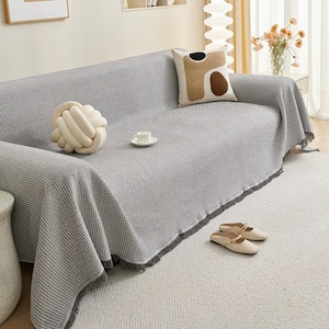 May include: A gray and white checkered sofa cover with fringe detail, draped over a gray sofa. A decorative knot pillow, a patterned pillow, and a white teacup and saucer are on the sofa. Beige slip-on shoes rest on the rug.