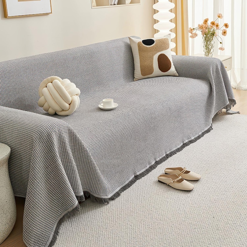 May include: A gray and white checkered sofa cover with fringe detail, draped over a gray sofa. A decorative knot pillow, a patterned pillow, and a white teacup and saucer are on the sofa. Beige slip-on shoes rest on the rug.