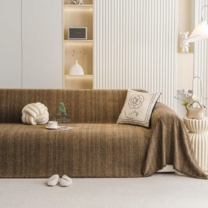 May include: A brown sofa with a textured herringbone pattern is covered with a matching throw blanket. A decorative pillow with a rose design and a white knot pillow are on the sofa. White slippers and a coffee cup are on the floor.