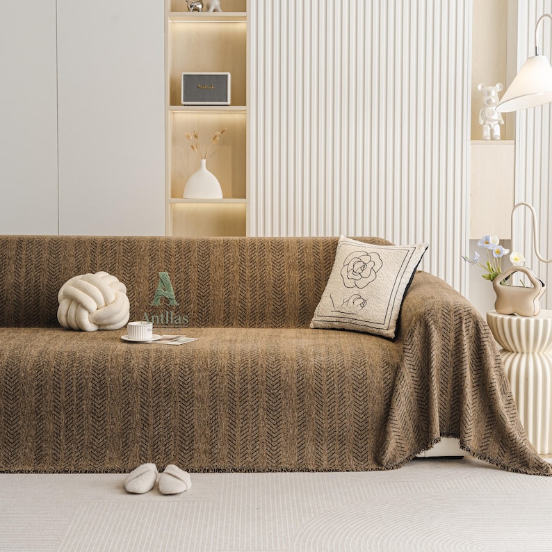 May include: A brown sofa with a textured herringbone pattern is covered with a matching throw blanket. A decorative pillow with a rose design and a white knot pillow are on the sofa. White slippers and a coffee cup are on the floor.