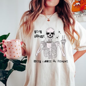 May include: A white t-shirt featuring a skeleton graphic holding a coffee cup. The text on the shirt reads "Dead inside, need coffee to revive."