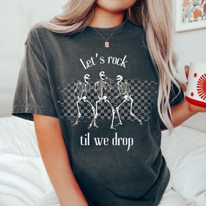 May include: A dark gray t-shirt with a graphic of three skeletons dancing and the text "Let's rock til we drop".