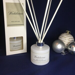 May include: A white reed diffuser with white reeds, a silver collar, and the text "Wickilicious" on the front. The box on the left also has the text "Wickilicious" and "Reed Diffuser". The fragrance is "Pomegranate Noir".