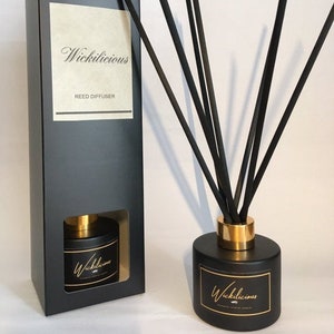 May include: A black reed diffuser set with a matching box. The diffuser has a black cylindrical base with a gold band and a label that reads "Wickilicious". Black reeds are inserted into the diffuser. The box also has the text "Wickilicious" and "Reed Diffuser".