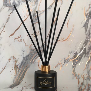 May include: A black diffuser bottle with black reeds and a gold collar. The bottle has a label that reads "Wickilicious" in gold script. The background is a marble pattern with gold veining.