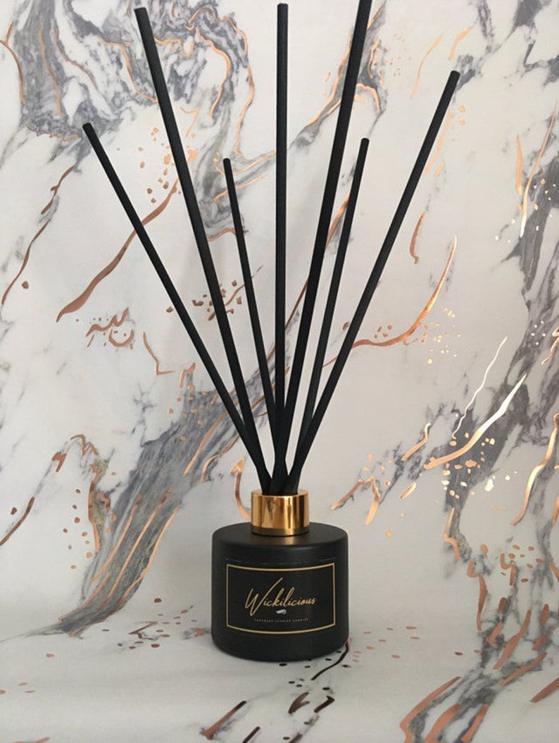 May include: A black diffuser bottle with black reeds and a gold collar. The bottle has a label that reads "Wickilicious" in gold script. The background is a marble pattern with gold veining.
