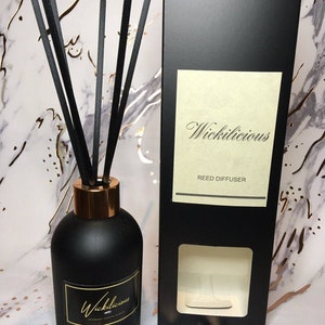 May include: A black reed diffuser with a gold-colored rim and black reeds. The diffuser has a label with the word "Wickilicious" in gold script. A black box with the same label is next to the diffuser.