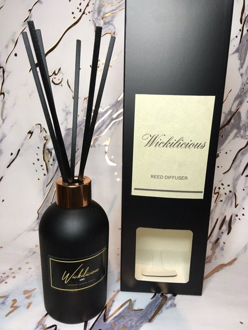 May include: A black reed diffuser with a gold-colored rim and black reeds. The diffuser has a label with the word "Wickilicious" in gold script. A black box with the same label is next to the diffuser.