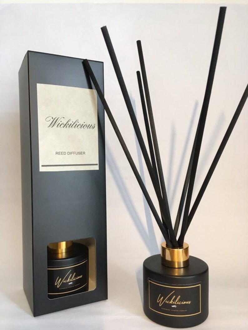 May include: A black reed diffuser set with a matching box. The diffuser has a black cylindrical base with a gold band and a label that reads "Wickilicious". Black reeds are inserted into the diffuser. The box also has the text "Wickilicious" and "Reed Diffuser".