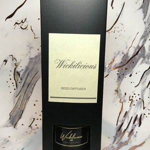 May include: A black rectangular box with the text "Wickilicious" in elegant script. The box contains a reed diffuser, with the product name also displayed on the diffuser itself. The background features a marble pattern with gold accents, creating a luxurious aesthetic.