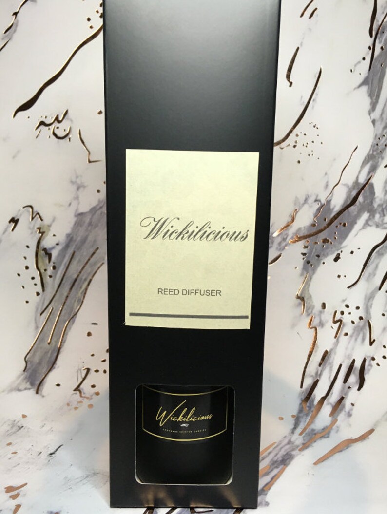 May include: A black rectangular box with the text "Wickilicious" in elegant script. The box contains a reed diffuser, with the product name also displayed on the diffuser itself. The background features a marble pattern with gold accents, creating a luxurious aesthetic.