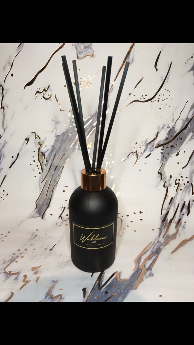 May include: A black diffuser bottle with a gold rim and black reeds. The bottle has a label with gold text that reads "Wickilicious". The background is a marble pattern with gold accents.