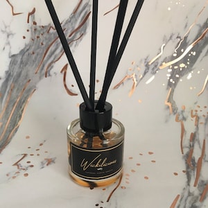 May include: A glass diffuser bottle with black reeds. The bottle has a black label with gold text reading "Wickilicious". The background is white with gold and grey marbling. The bottle contains a golden liquid.