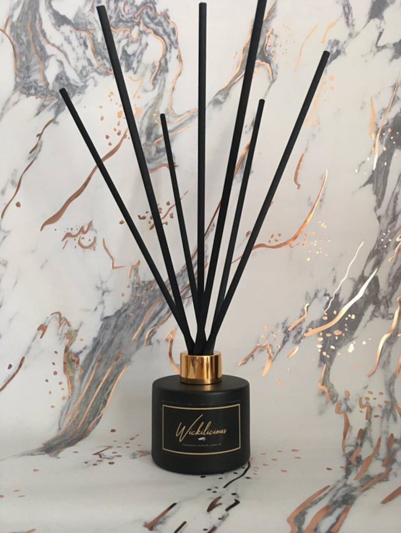 May include: A black diffuser bottle with a gold collar and black reeds. The bottle has a label that reads "Wickilicious". The background is a marble pattern with gold accents. A home fragrance product.