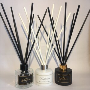 May include: Three reed diffusers with black and white reeds. The diffusers are in clear, white, and black glass bottles. Each bottle has a label with the word "Wickilicious" in gold lettering. The clear bottle has a black cap, the white bottle has a silver cap, and the black bottle has a gold cap.