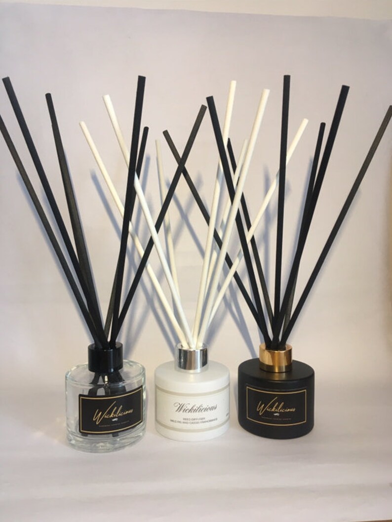 May include: Three reed diffusers with black and white reeds. The diffusers are in clear, white, and black glass bottles. Each bottle has a label with the word "Wickilicious" in gold lettering. The clear bottle has a black cap, the white bottle has a silver cap, and the black bottle has a gold cap.