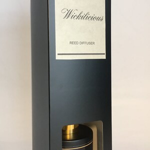 May include: Black rectangular box with a window revealing a black reed diffuser. The diffuser has a gold top and a black label with gold script that reads "Wickilicious". The box has a label that reads "Wickilicious REED DIFFUSER".