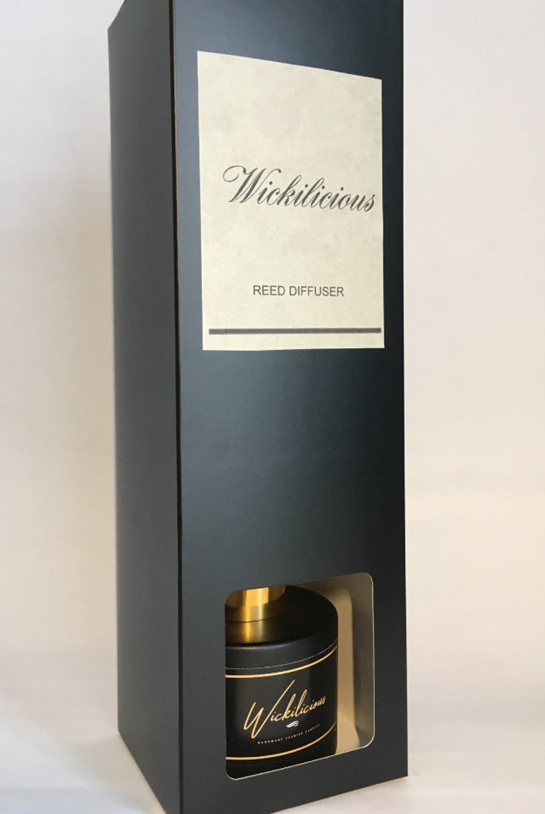 May include: Black rectangular box with a window revealing a black reed diffuser. The diffuser has a gold top and a black label with gold script that reads "Wickilicious". The box has a label that reads "Wickilicious REED DIFFUSER".