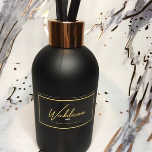 May include: A black diffuser bottle with a copper-colored collar and black reeds. The bottle has a label with the word "Wickilicious" in gold script. The background is a marble pattern with gold accents.
