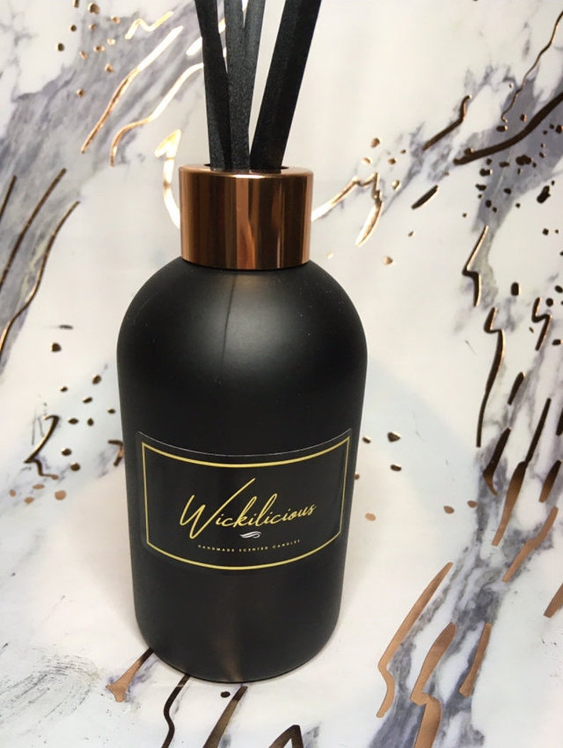 May include: A black diffuser bottle with a copper-colored collar and black reeds. The bottle has a label with the word "Wickilicious" in gold script. The background is a marble pattern with gold accents.