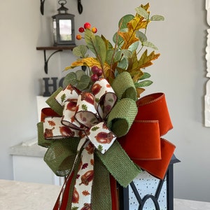 May include: A black lantern decorated with a fall-themed bow and foliage. The bow features a burlap-style green ribbon, a rust-colored ribbon, and a ribbon printed with acorns and leaves. The arrangement includes faux leaves, berries, and greenery.