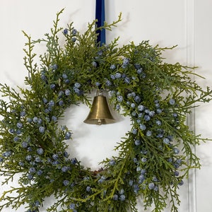 May include: A circular wreath made of green cedar branches and clusters of small, frosted blue berries. A polished brass bell hangs from the center of the wreath, suspended by a dark blue velvet ribbon that extends upward from the top.