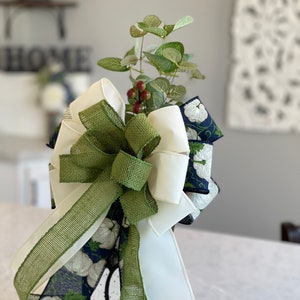 K&ouml;nnte beinhalten: Eine dekorative Schleife mit einer Mischung aus Texturen und Farben. Die Schleife enth&auml;lt wei&szlig;en Satin, gr&uuml;nes Jute und marineblaues Band mit wei&szlig;en Blumenmustern. Sie ist mit k&uuml;nstlichem Gr&uuml;n und roten Beeren verziert, geeignet f&uuml;r die Inneneinrichtung.