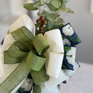 K&ouml;nnte beinhalten: Eine dekorative Schleife mit wei&szlig;en, gr&uuml;nen und marineblauen B&auml;ndern. Die Schleife ist mit k&uuml;nstlichem Gr&uuml;n geschm&uuml;ckt, darunter Eukalyptusbl&auml;tter und rote Beeren. Das marineblaue Band hat ein wei&szlig;es Blumenmuster.