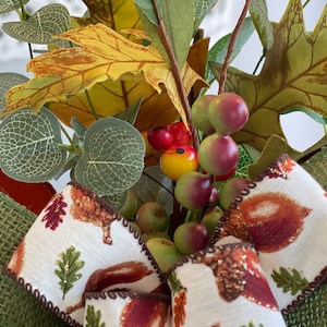May include: A close-up of a fall-themed decorative wreath. The wreath features a variety of faux foliage, including green, yellow, and brown leaves, along with small red and green berries. A cream-colored ribbon with acorn and leaf designs adds a decorative touch.