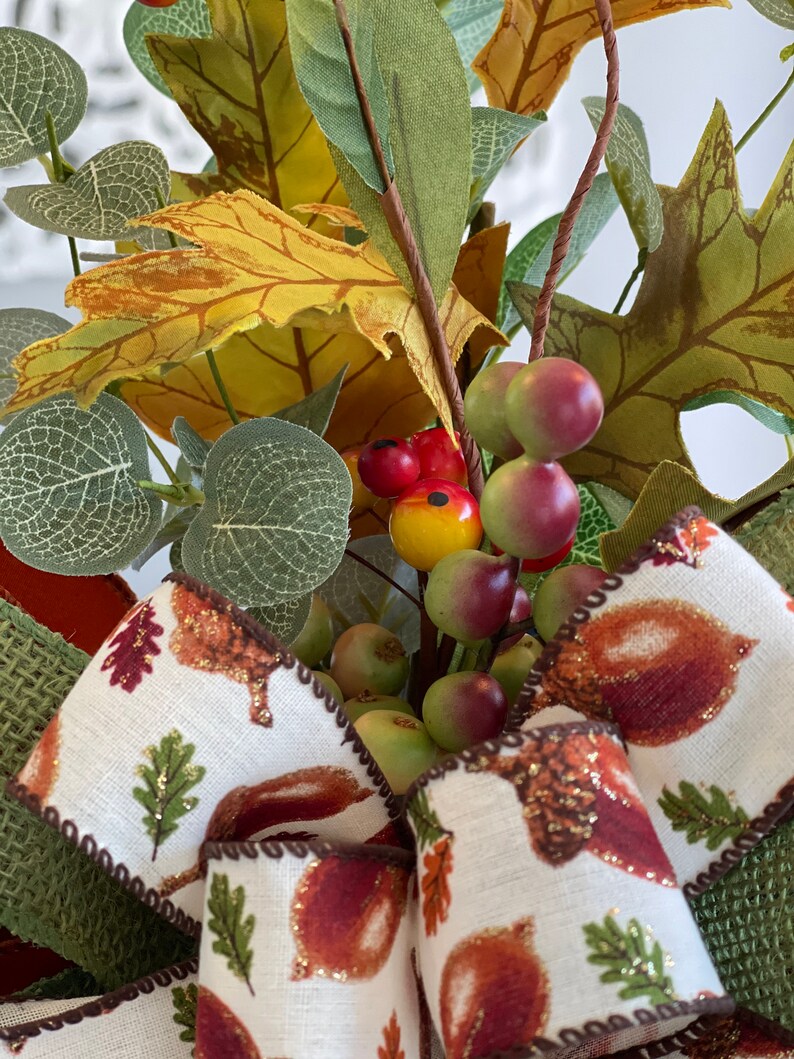 May include: A close-up of a fall-themed decorative wreath. The wreath features a variety of faux foliage, including green, yellow, and brown leaves, along with small red and green berries. A cream-colored ribbon with acorn and leaf designs adds a decorative touch.